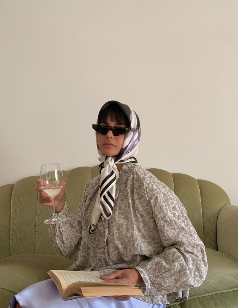 Woman drinking wine and reading a book with a scarf over her head.