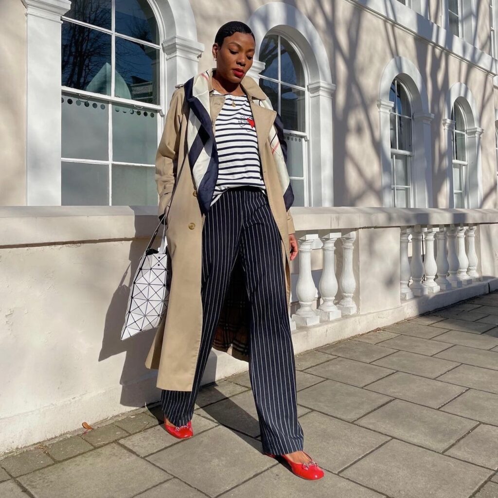 woman wearing black trousers, striped t-shirt, trench coat, and rad ballet flats