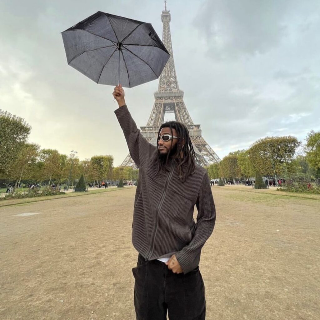 man wearing a corduroy jacket and cargo pants while holding up an umbrella in front of the Eiffel Tower