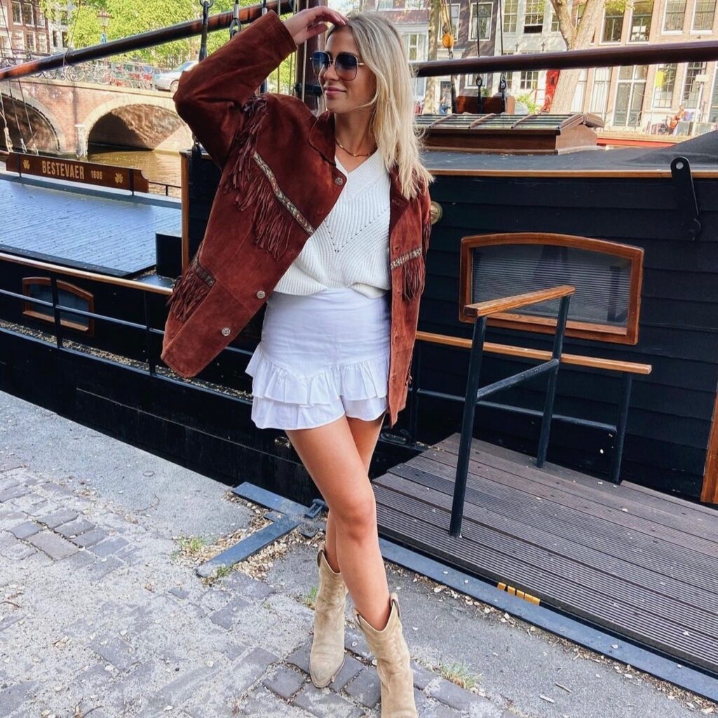 woman in brown fringe jacket with off-white sweater, white skirt, and cowboy boots