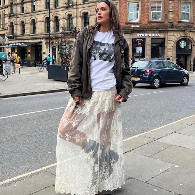 woman wearing long cream-colored lace skirt with black cowboy boots, graphic tee, and khaki bomber jacket