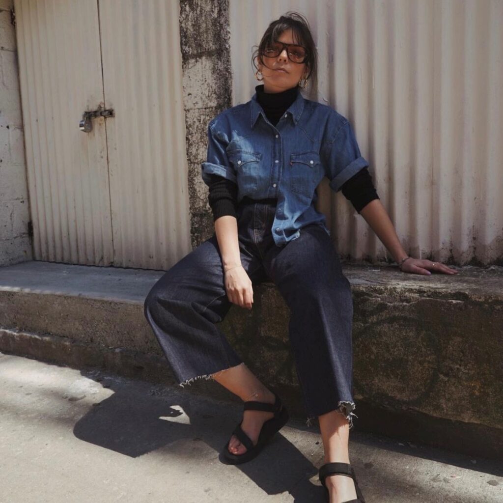 woman sitting outside wearing a black turtleneck, denim shirt, cropped jeans, and black sandals