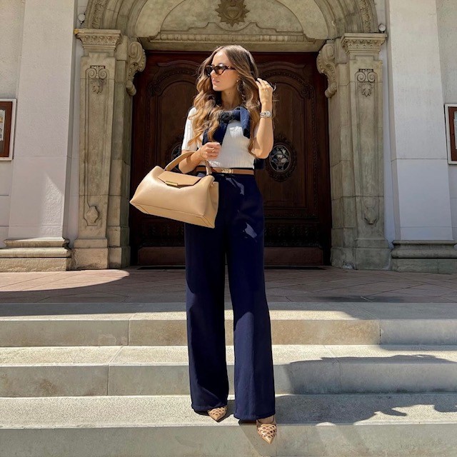 woman in navy blue pants and cropped white sweater with tan handbag