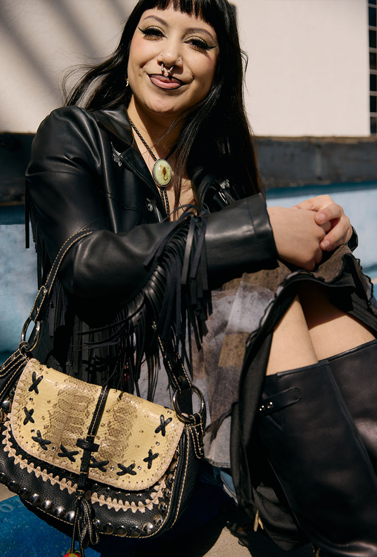 female model wearing a black fringe jacket midi skirt and tall black boots holding a snakeskin purse