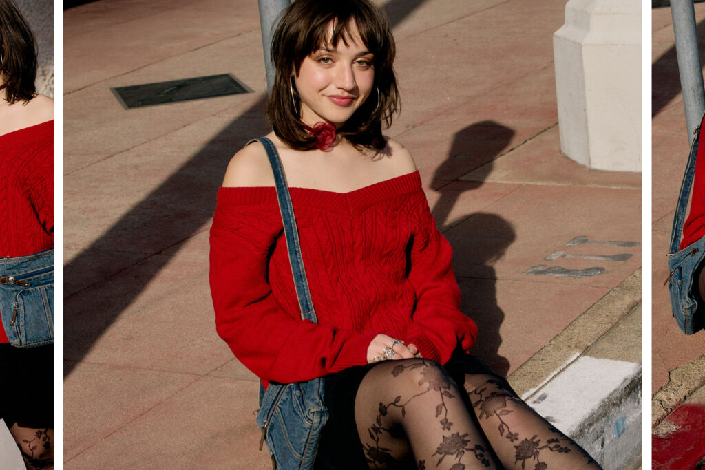 Emily wearing a red off the shoulder sweater, black skirt, floral tights sitting on the curb