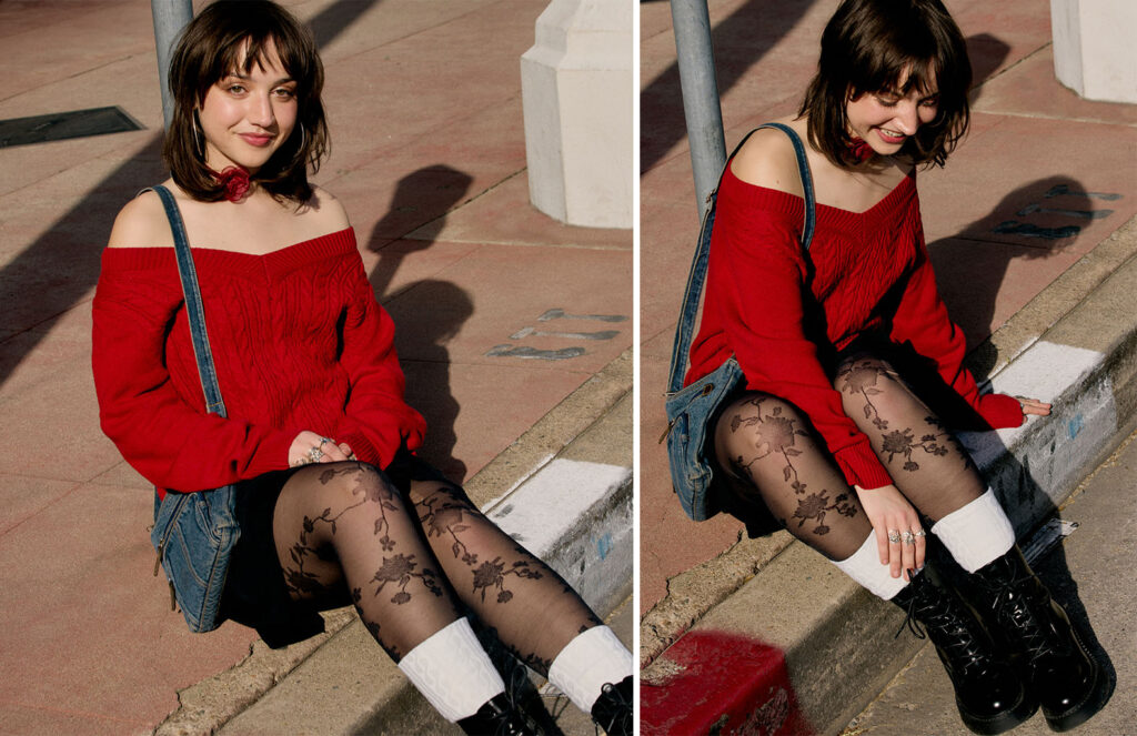 Emily wearing a red off the shoulder sweater, black skirt, floral tights sitting on the curb