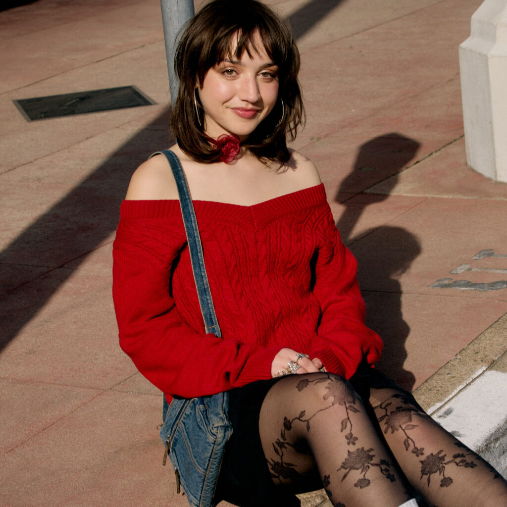 Emily wearing a red off the shoulder sweater, black skirt, floral tights sitting on the curb