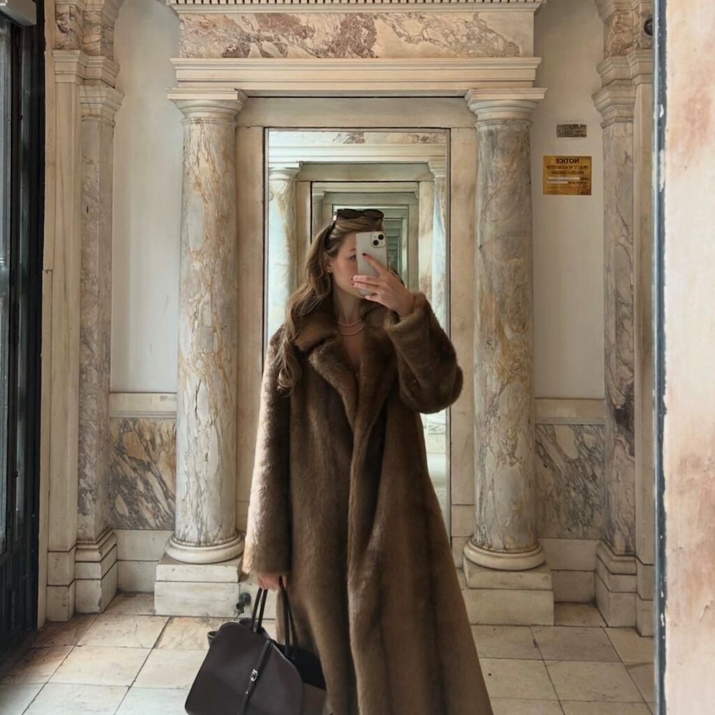 woman taking selfie in a mirror wearing a long brown faux fur coat 