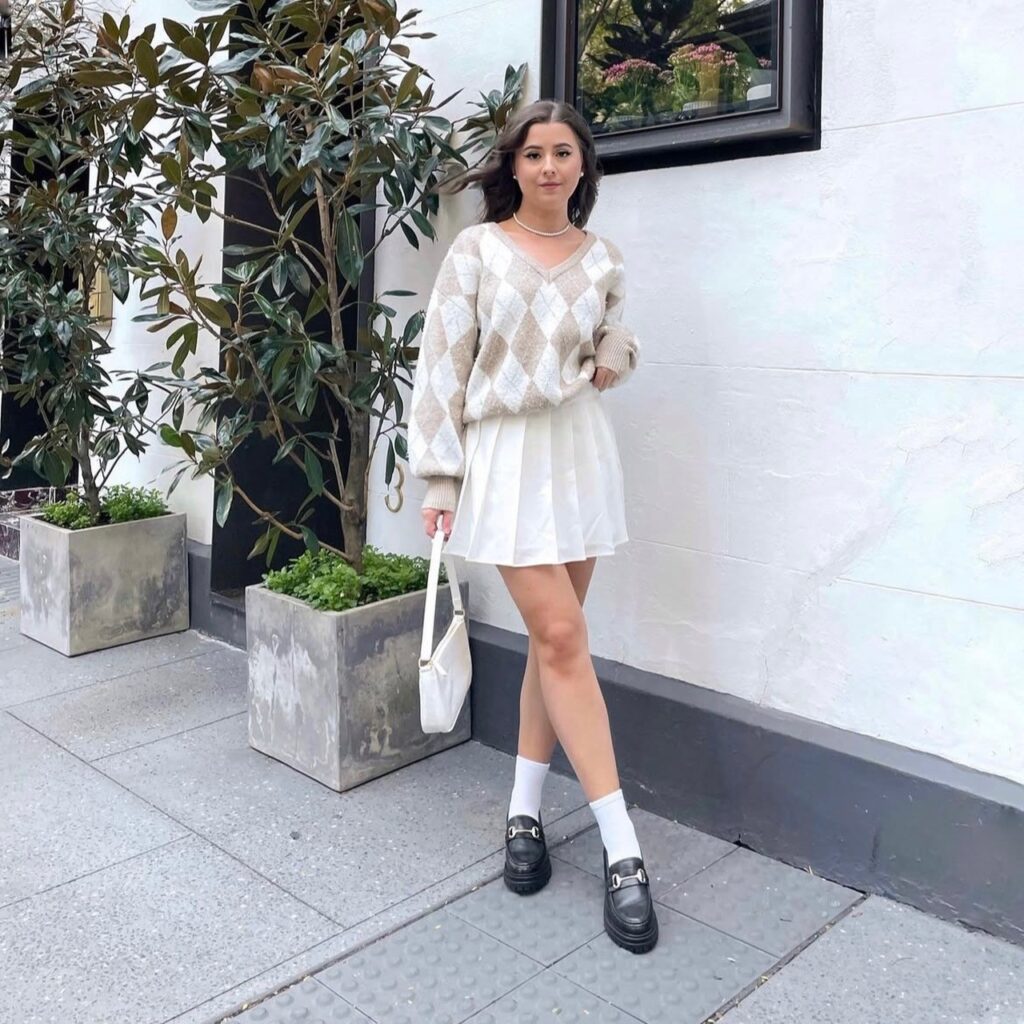 woman in white pleated skirt, argyle sweater, and black platform loafers