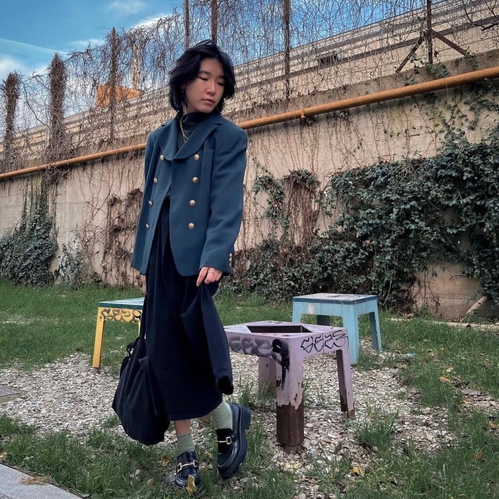 person in long black dress, gray blazer, mint green socks, and black loafers