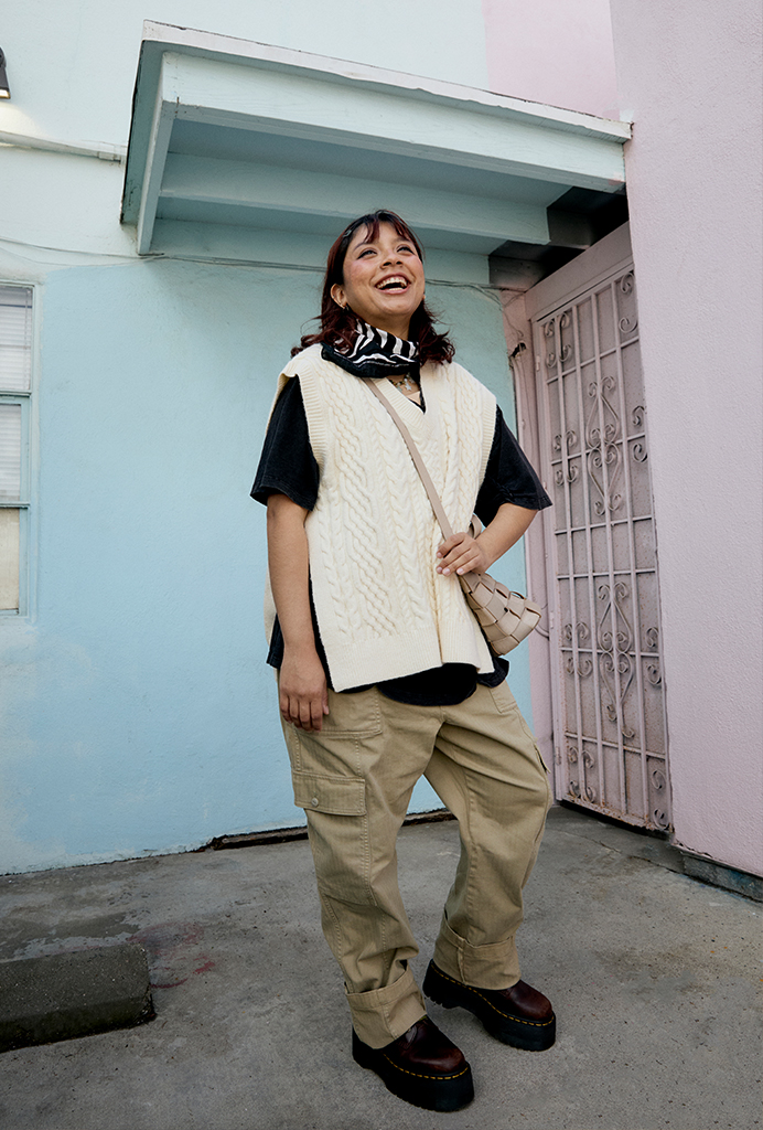 Lee wearing a black tee, cream knit vest on top, black bandanna tied around their neck, khaki cargo pants, brown docs smiling at the camera