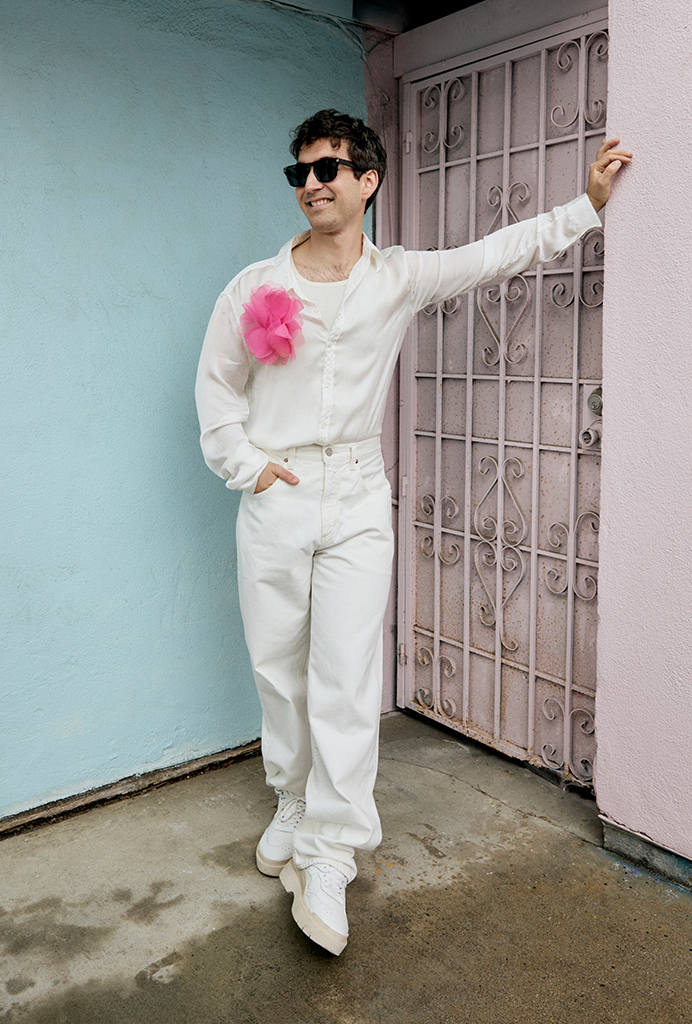 Charlie leaning on a pink wall wearing a white button up shirt, white pants and white shoes with a pink flower pinned to his shirt