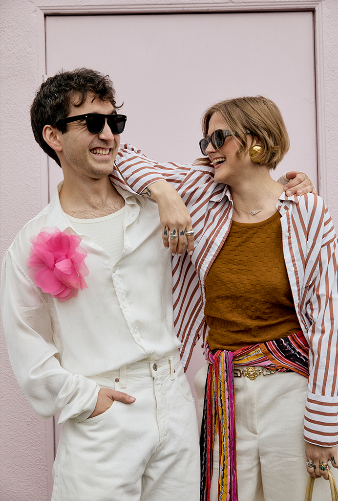 Charlie wearing a white button up and white pants with a pink flower pinned to his shirt Britt posed next to Charlie wearing a white and orange striped long sleeve button up, brown tank top under white shorts and a colorful striped longsleeve tied around her waist