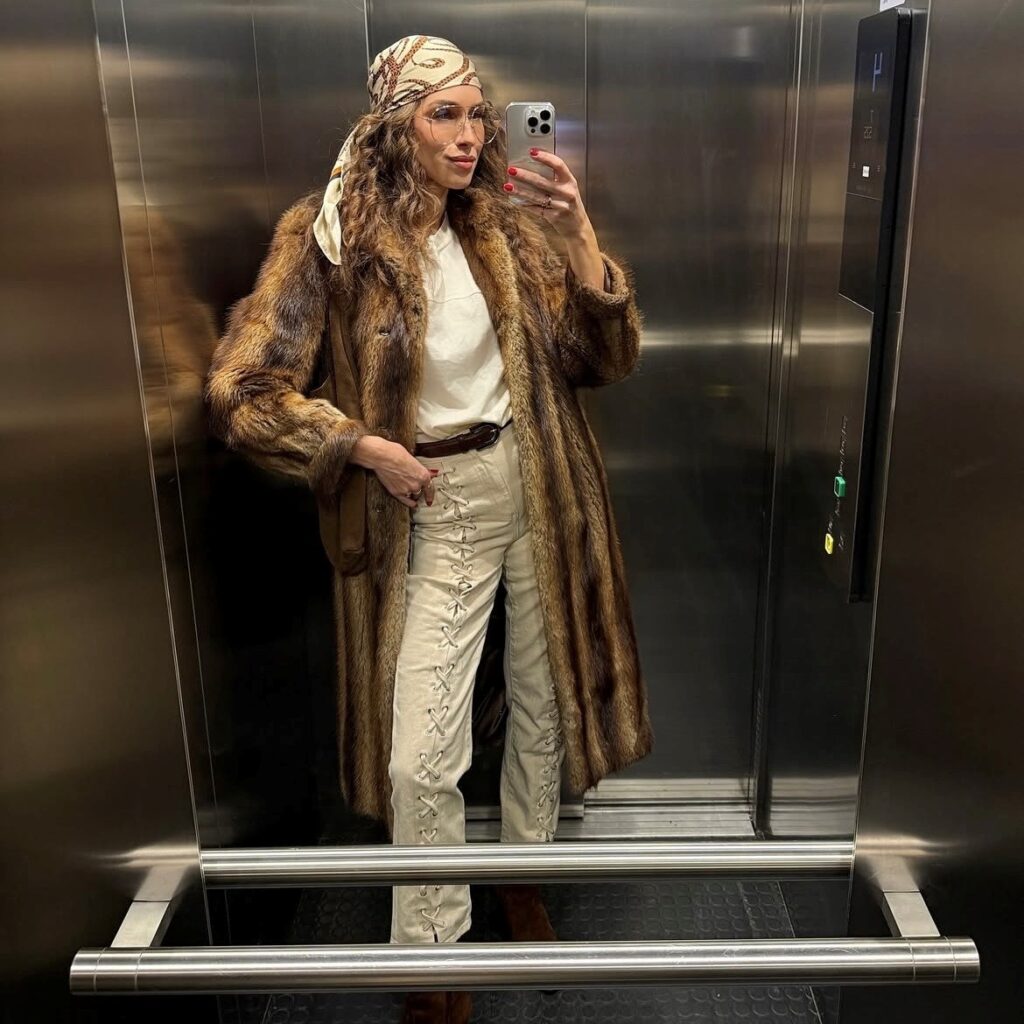 woman taking selfie in elevator wearing a fur coat with silk scarf tied over hair