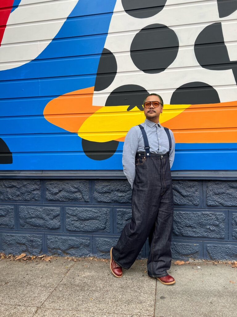 Arnel in high-waisted dark denim pants with suspenders and blue button-down