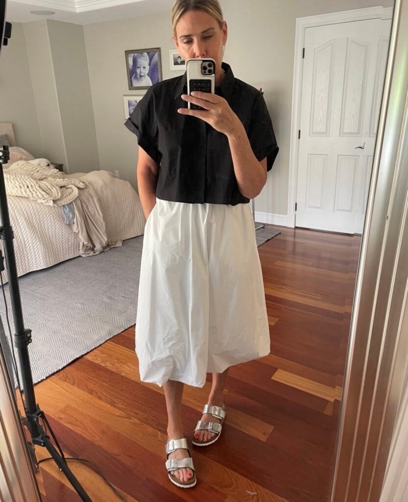woman taking a selfie in white skirt with black blouse and silver sandals