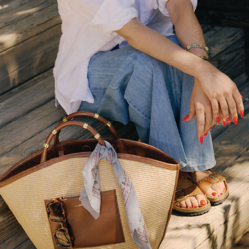 Accessory photo featuring a model sitting on a staircase wearing blue wide leg denim, a white linen blouse, red nails, brown birkenstock shoes, and a large woven tote bag