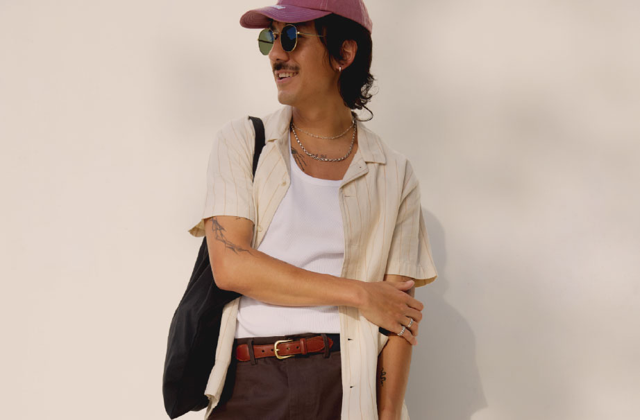 male model wearing a red hat, sunglasses, cream button up open over a white tank top, brown pants and brown belt, holding a black canvas tote bag