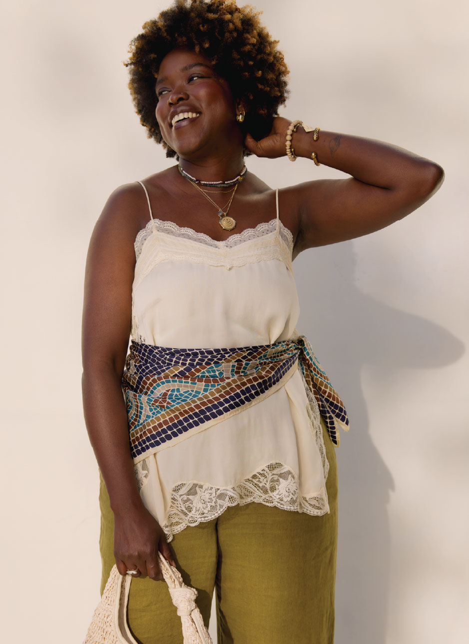 female model wearing a white lace camisole with a blue mosaic pattern scarf across her waist, green linen pants, holding a woven purse