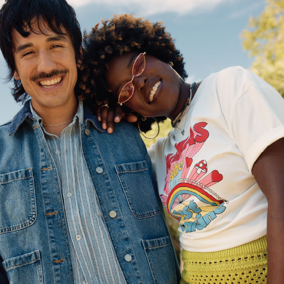 two models smiling together; left male model wearing med wash denim jacket over a blue stripe button up, right female model wearing pink sunglasses, white rainbow graphic tee, and green crochet skirt