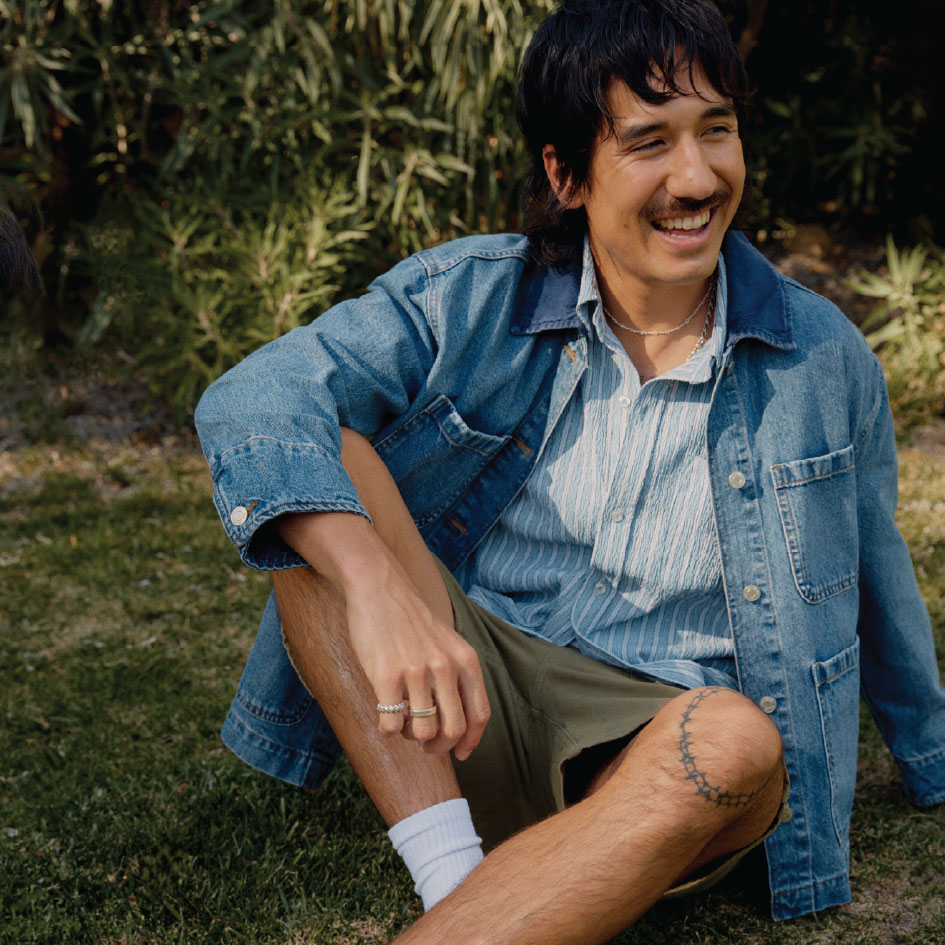 male model sitting on grass wearing a med wash denim jacket over a light blue stripe button up and green shorts