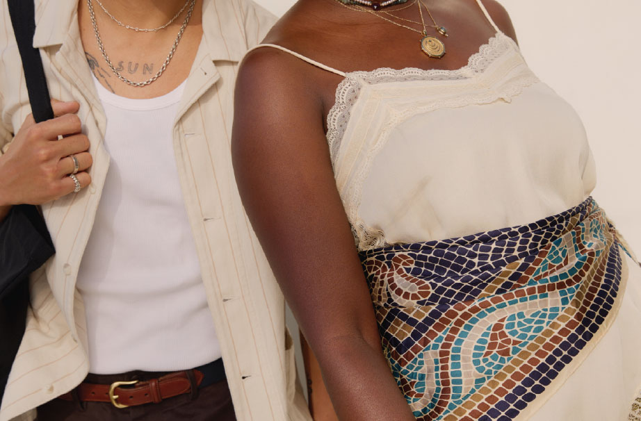 outfit detail shot of two models; left male model is wearing a cream button up open over a white tank top with brown pants and a brown leather belt, right female model is wearing a cream lace camisole with a blue mosaic patterned scarf tied across her waist