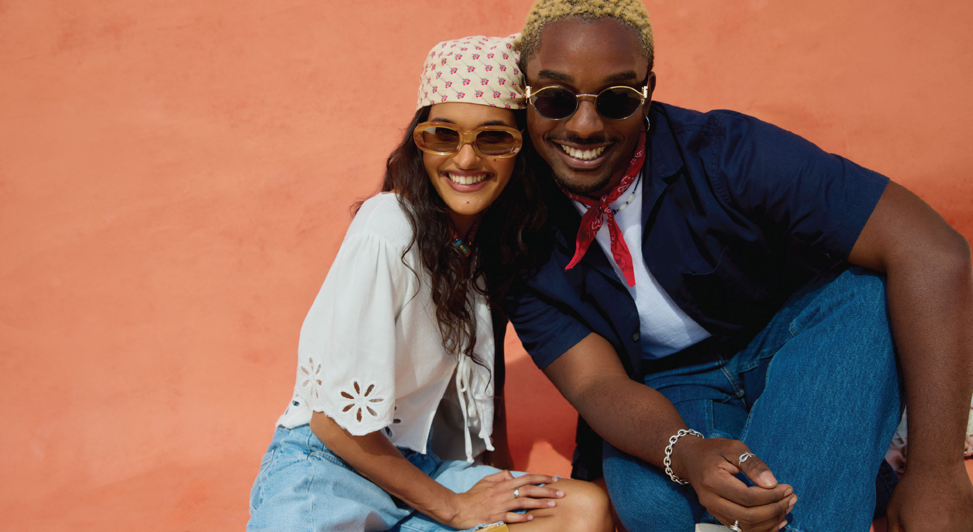 two models sitting smiling together; left female model is wearing a white patterned head scarf, sunglasses, a white linen top, and blue denim shorts, right male model is wearing sunglasses, a red neck scarf, navy linen button up over a white tee and med wash denim