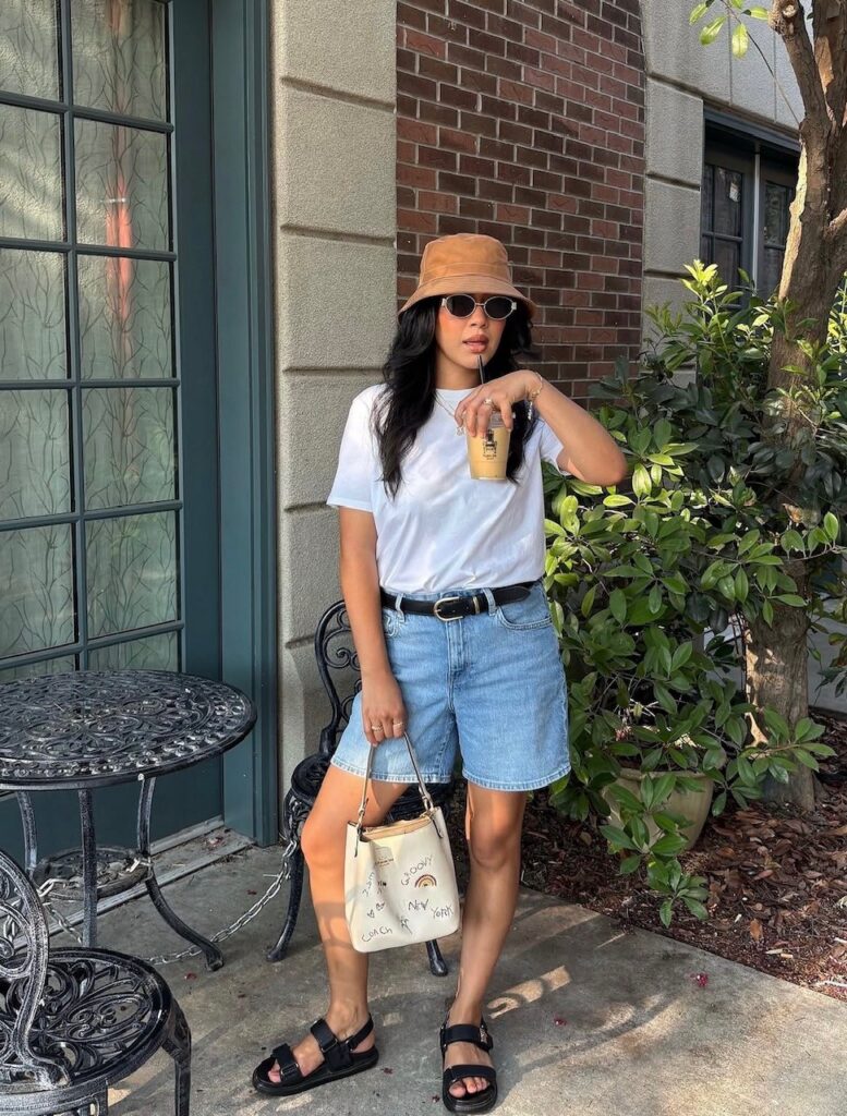 Woman wearing denim shorts, white tee, and black sporty sandals with a bucket hat.