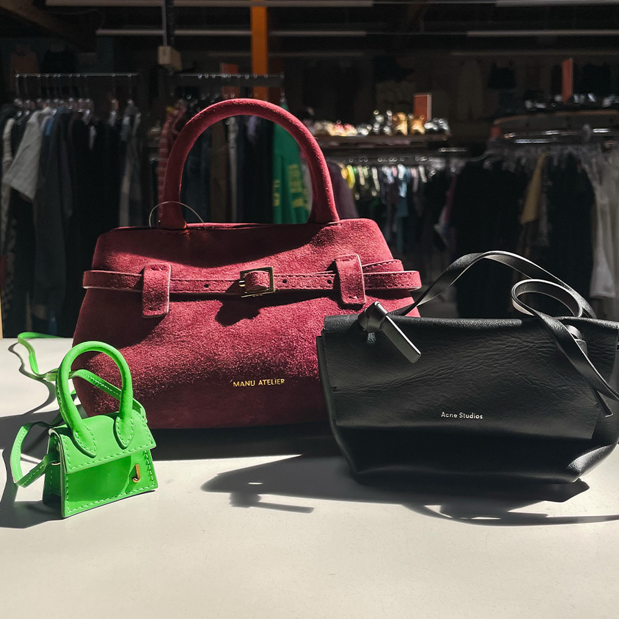 Three designer handbags displayed on a counter in sunlight: a burgundy suede Manu Atelier bag with a top handle and belt detail, a small bright green micro bag with a chain strap, and a black leather Acne Studios crossbody bag.