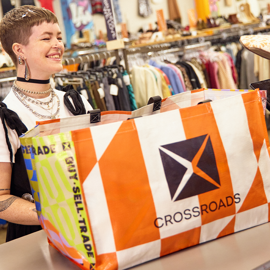 A smiling person with a short pixie cut, layered necklaces, and multiple piercings stands at a Crossroads counter, dropping off clothes in a large orange-and-white reusable tote bag with the Crossroads logo and "Buy. Sell. Trade." printed on the side.