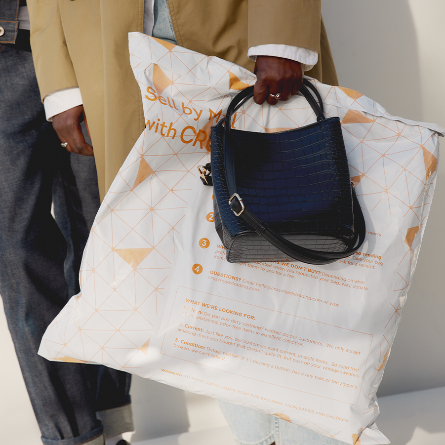 A person wearing a tan coat holds a large white "Sell by Mail with Crossroads" shipping bag printed with instructions and guidelines for mailing in clothes.