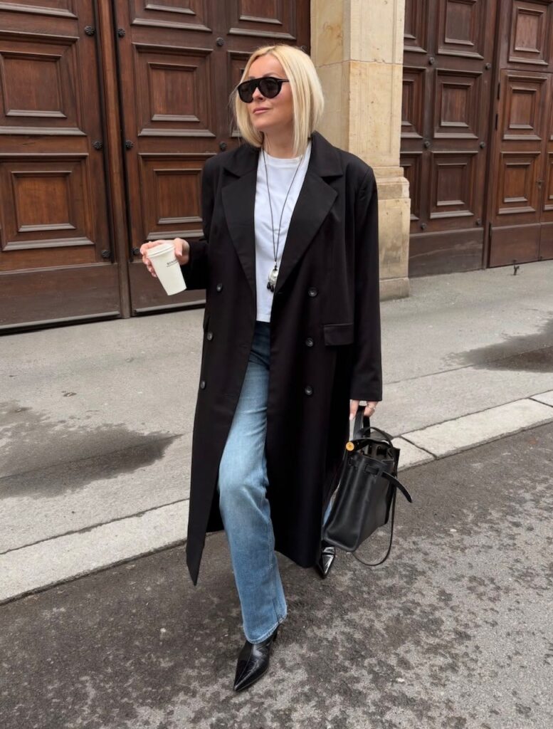 woman in jeans, white tee, and long black trench, with black pointed boots